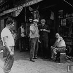 Men sitting and standing outside of shops along Brooklyn waterfront.